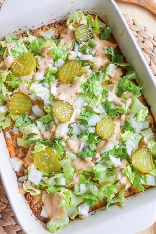 A casserole dish filled with a layered salad. The top layer includes chopped lettuce, sliced pickles, diced onions, and drizzled dressing over a visible layer of cheese and ground meat. The dish rests on a woven mat.