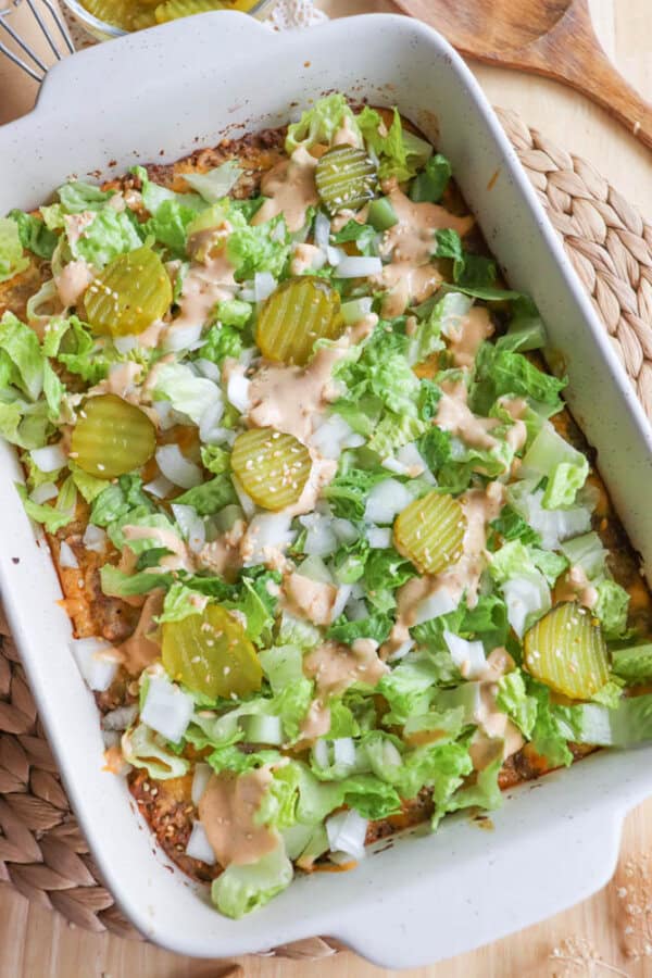 A casserole topped with diced lettuce, pickles, chopped onions, and a drizzle of sauce. The dish is in a rectangular white baking dish on a woven mat, with a wooden spoon in the background.
