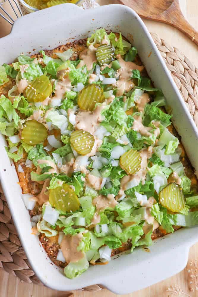 A casserole topped with diced lettuce, pickles, chopped onions, and a drizzle of sauce. The dish is in a rectangular white baking dish on a woven mat, with a wooden spoon in the background.