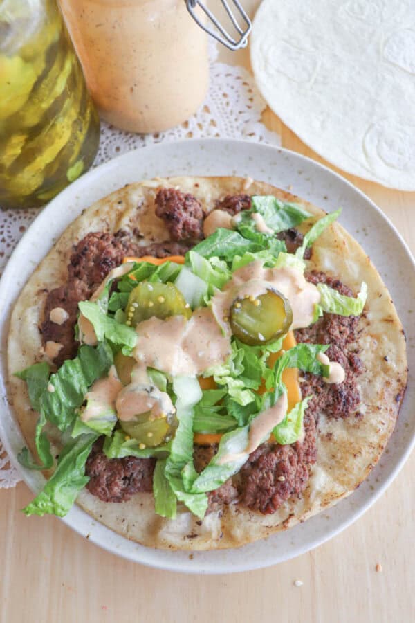 A plate with a flatbread topped with ground meat, lettuce, pickles, and a creamy sauce. A jar of pickles, sauce, and a round tortilla are visible in the background on a wooden table with a lace doily.