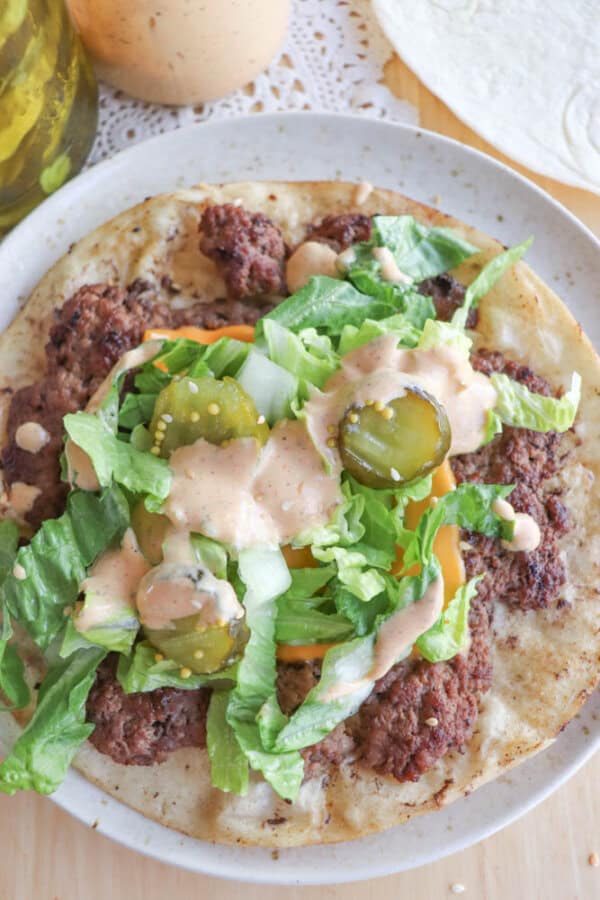 A plate with a tortilla topped with cooked ground beef, shredded lettuce, sliced pickles, melted cheese, and a drizzle of creamy sauce. The dish is garnished with herbs and surrounded by a jar of pickles and a doily.