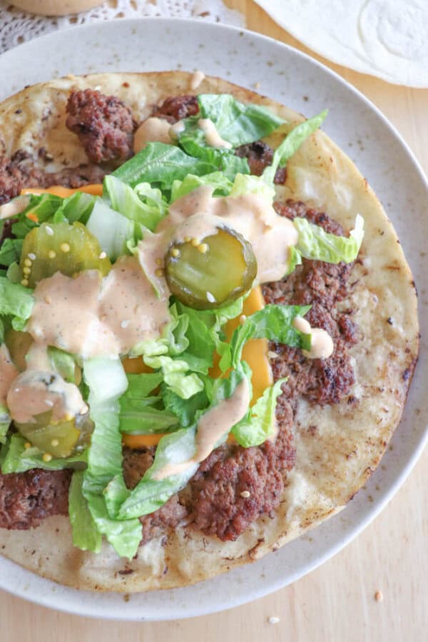 A plate with a tortilla topped with seasoned ground beef, shredded lettuce, pickles, shredded cheese, and drizzled with a creamy sauce. The dish is placed on a light wooden table.