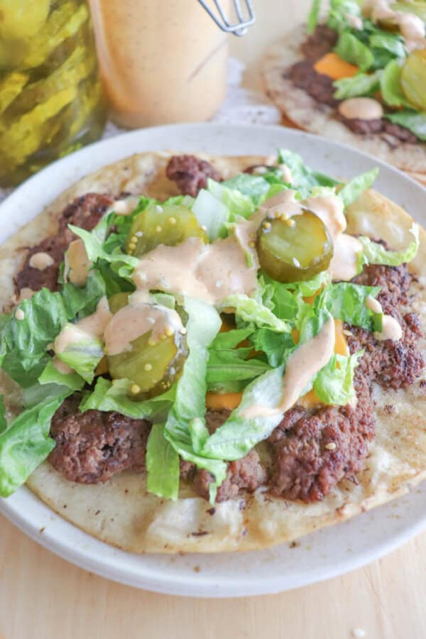 A flatbread topped with beef pieces, shredded lettuce, pickles, and drizzled with creamy sauce. A jar of pickles and a container of sauce are in the background.