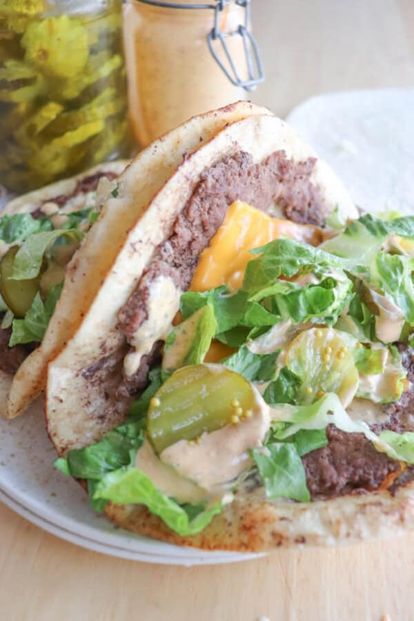 A close-up of a taco filled with ground beef, melted cheddar cheese, sliced pickles, shredded lettuce, and a creamy sauce. A jar of pickles is in the background on a wooden surface.