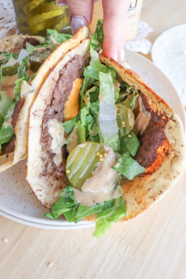 A hand holds a tortilla filled with ground beef, melted cheese, lettuce, pickles, and a creamy sauce. A jar of pickles is visible in the background on a wooden surface.