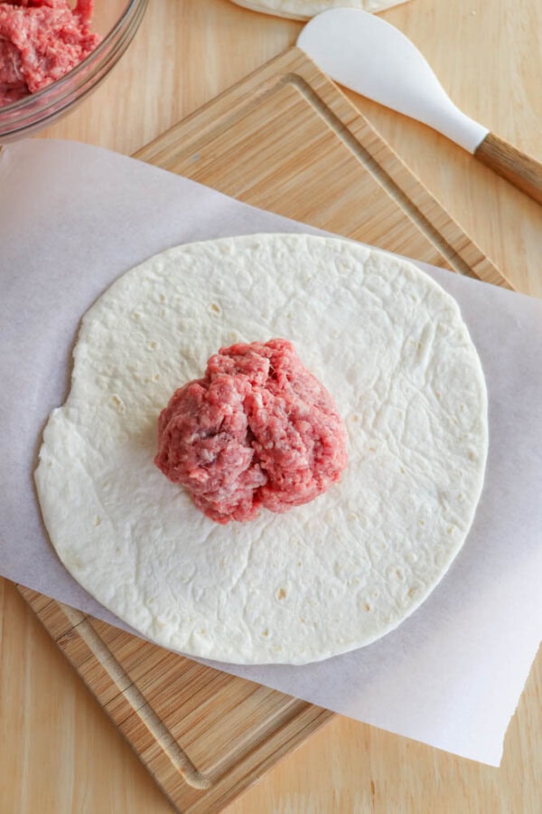 A tortilla on a wooden board with a sheet of parchment paper underneath. Raw ground meat is placed at the center of the tortilla. A white spatula and a bowl of more ground meat are visible in the background.