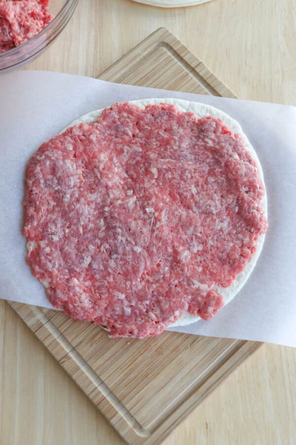 Raw ground meat spread evenly over a round pizza dough on parchment paper, placed on a wooden cutting board. A portion of a mixing bowl is visible in the upper corner.