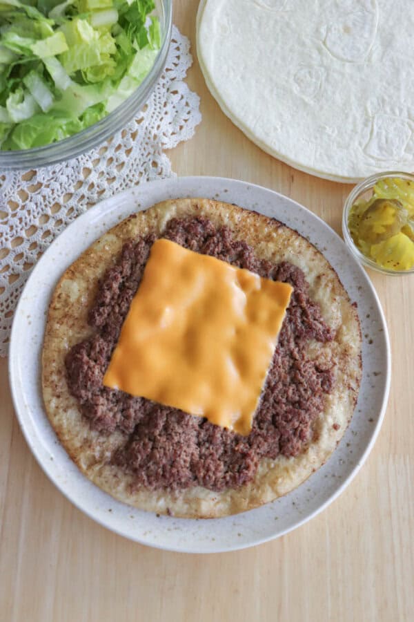 A plate with a flatbread topped with ground meat and a slice of American cheese. Nearby are a bowl of shredded lettuce, a stack of tortillas, and a small bowl of pickles on a wooden surface with a lace doily.