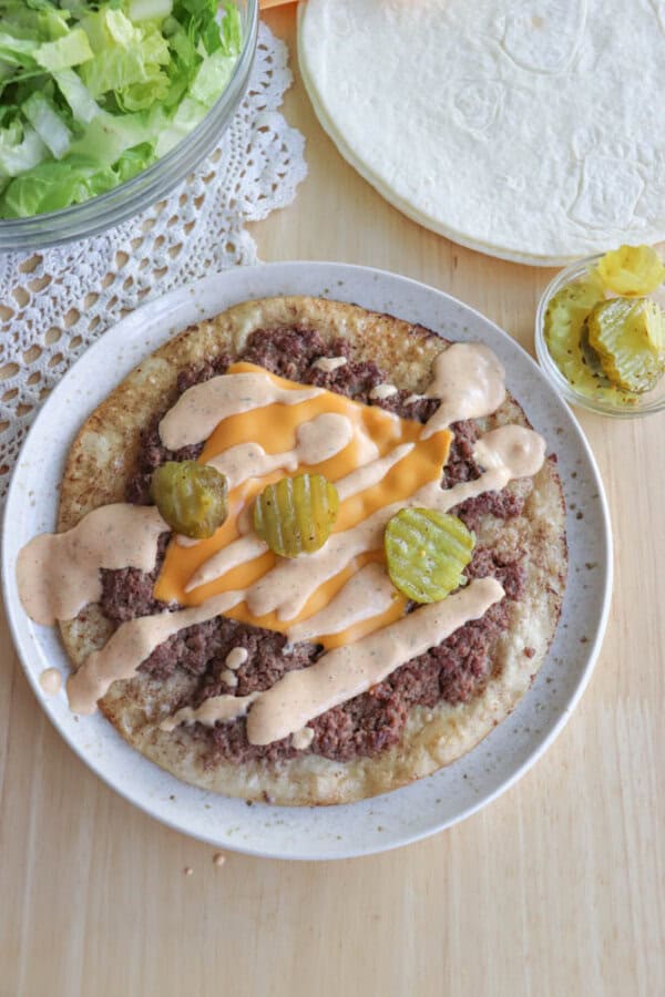 A flat taco with ground meat, melted cheese, pickles, and sauce drizzled on top sits on a speckled plate. A bowl of lettuce, a stack of tortillas, and a small bowl of sliced pickles are nearby on a wooden surface with a lace doily.