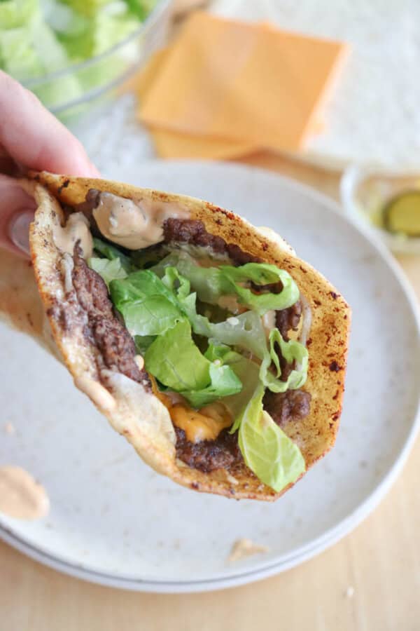 Close-up of a hand holding a taco filled with refried beans, lettuce, melted cheese, and sauce in a crispy shell. A plate with taco ingredients such as lettuce and cheese is blurred in the background.