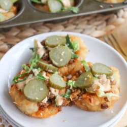 Two loaded muffins on a white plate feature a savory filling topped with sliced pickles, shredded lettuce, and drizzles of sauce. A muffin tray with more muffins is in the background. The setting is on a woven mat with a lace doily nearby.