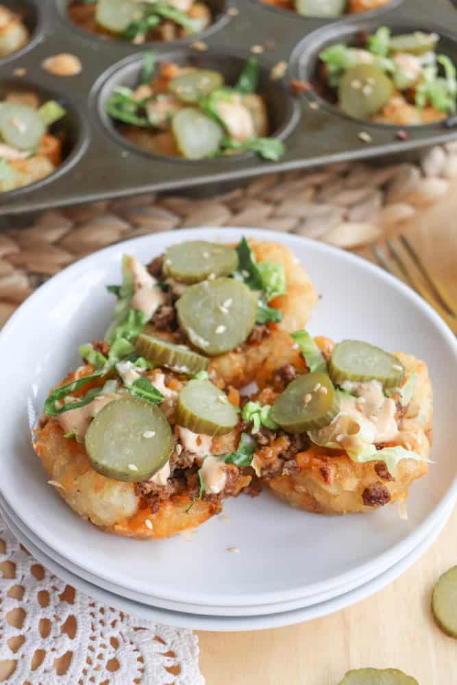 Two loaded muffins on a white plate feature a savory filling topped with sliced pickles, shredded lettuce, and drizzles of sauce. A muffin tray with more muffins is in the background. The setting is on a woven mat with a lace doily nearby.
