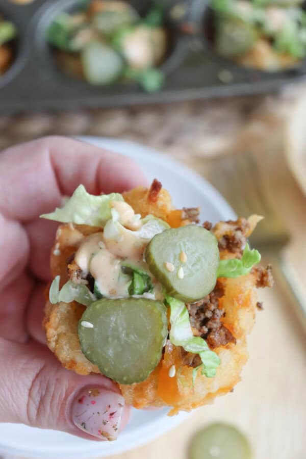 Close-up of a hand holding a tater tot cup filled with ground beef, melted cheese, sliced pickles, lettuce, and drizzled with sauce. Thereโs a sesame seed on top, with a muffin tray of similar snacks blurred in the background.