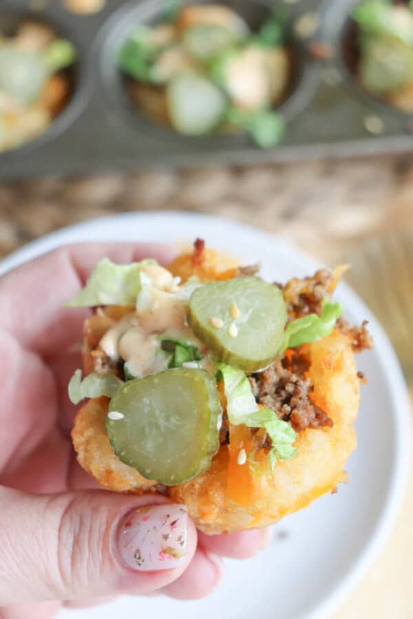 A hand holding a small, round bite-sized piece of food made with ground beef, lettuce, pickles, and sauce on a crispy base, likely a tater tot cup. A muffin tray with more of these is blurred in the background.