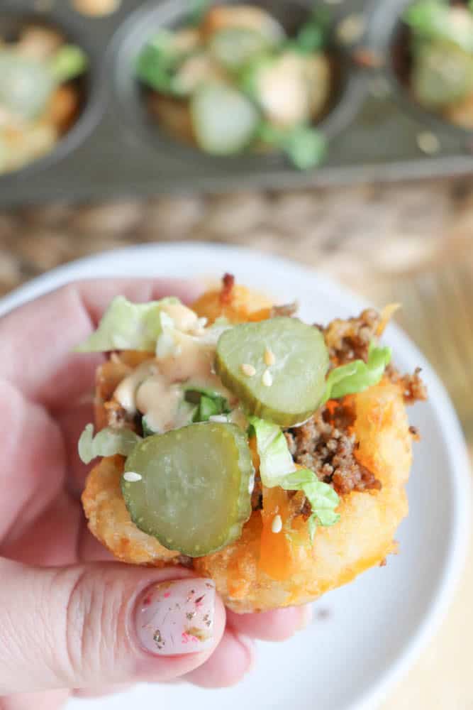 A hand holding a small, round bite-sized piece of food made with ground beef, lettuce, pickles, and sauce on a crispy base, likely a tater tot cup. A muffin tray with more of these is blurred in the background.
