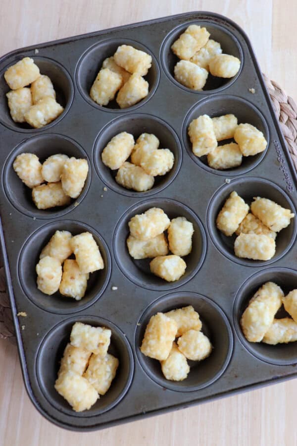 A muffin tin filled with tater tots, placed on a wooden surface. Each compartment contains a few golden-brown tater tots.