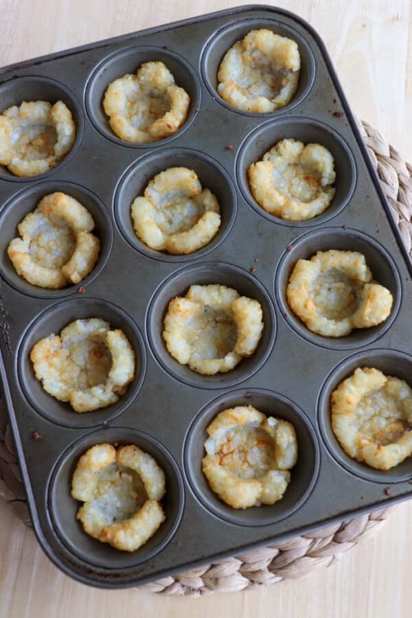 A muffin tray containing 12 crispy, golden-brown tater tots, each molded into cup shapes. The tray is placed on a woven mat, and the background is a light wooden surface.