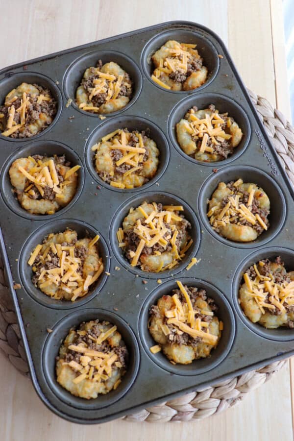 Muffin tin filled with unbaked hash brown cups, topped with ground meat and shredded cheese, on a wooden surface.