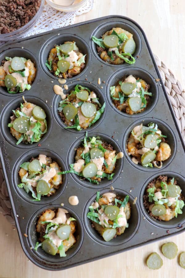 A muffin tin filled with taco cups, each topped with lettuce, sauce, and sliced pickles. The cups appear to have a mixture of meat and cheese. A small bowl with ingredients is visible in the background.