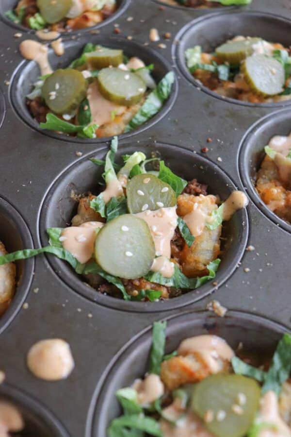 Close-up of a muffin tin containing savory mini dishes, resembling cheeseburger cups. They are filled with ground meat, shredded lettuce, pickles, and drizzled with a creamy sauce. Sesame seeds are sprinkled on top.