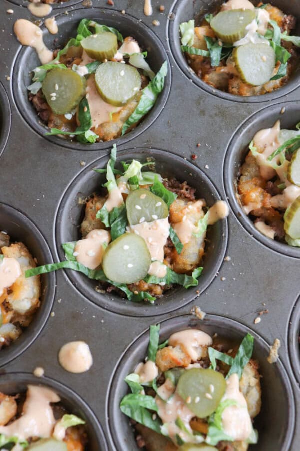 Mini savory cups in a muffin tin, topped with sliced pickles, shredded lettuce, sauce, and sesame seeds. The cups appear to contain a mixture of baked ingredients, possibly hash browns or a similar base. Some sauce is drizzled around the tin.