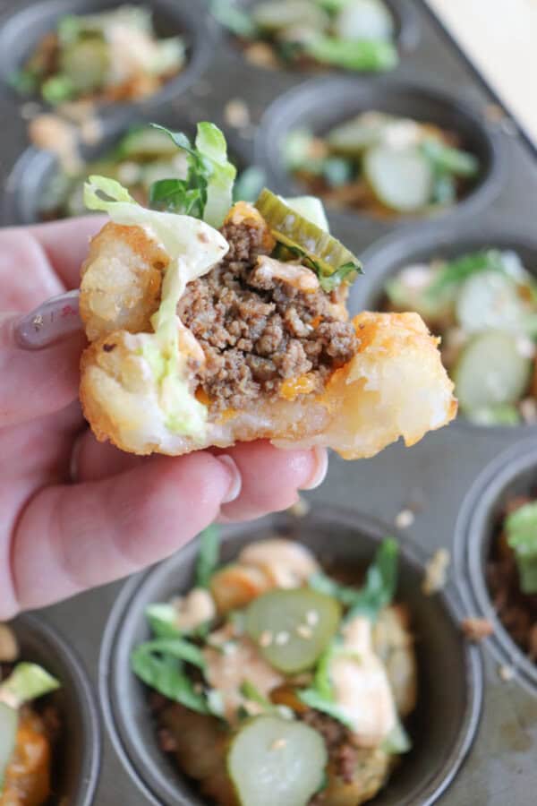 Close-up of a hand holding a mini burger cup made from tater tots, filled with ground beef, lettuce, and topped with pickles. A muffin tin with more prepared burger cups is visible in the background.