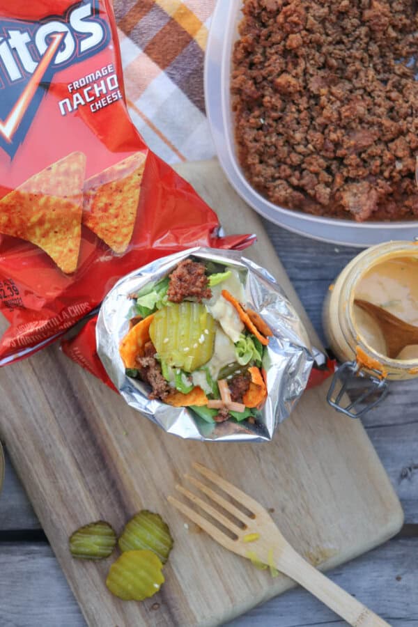 An open bag of Nacho Cheese Doritos filled with ground meat, lettuce, pickles, and sauce sits on a wooden board next to a fork, pickle slices, a bowl of ground meat, and a jar of sauce.