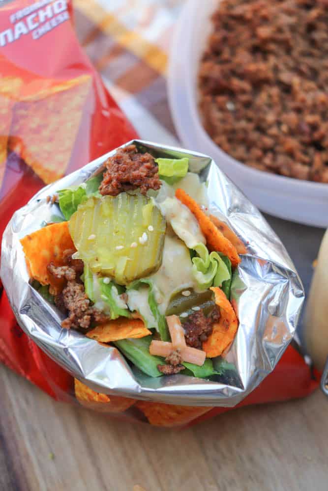 An open bag of nacho cheese chips filled with taco ingredients, including ground beef, lettuce, shredded cheese, pickles, and sauce, resembling a walking taco, with more meat in a container nearby.