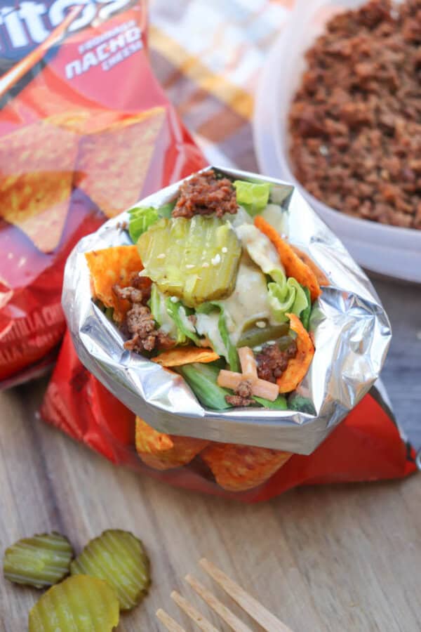 An open Doritos chip bag filled with taco meat, lettuce, cheese, and pickles, with a bag of chips and a container of taco meat in the background. Sliced pickles and a wooden fork are on the table nearby.