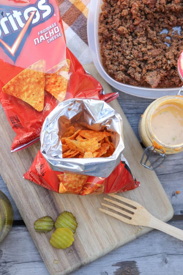 Open bag of Doritos nacho cheese chips on a wooden board with pickle slices, a wooden fork, a container of cooked ground beef, and a jar of creamy sauce nearby.