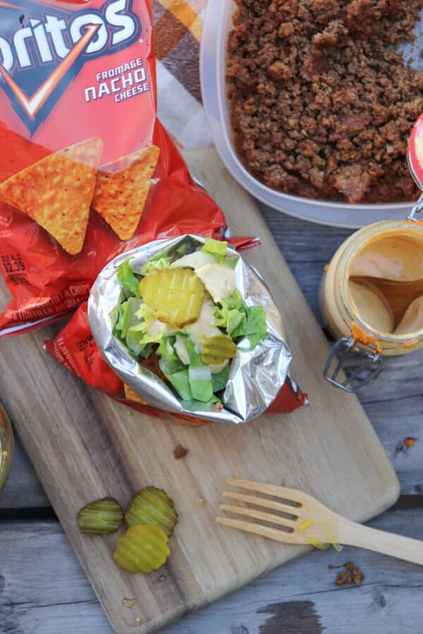 An open bag of Doritos filled with ground beef, lettuce, pickles, and sauce sits on a wooden board, with a jar of sauce, a container of cooked meat, and extra pickles nearby.