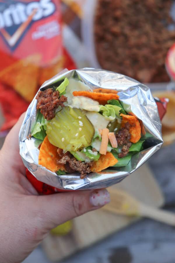 A hand holding a foil-wrapped taco filled with ground beef, lettuce, shredded cheese, pickles, and crunchy Fritos chips. An open bag of Fritos and taco ingredients are blurred in the background.