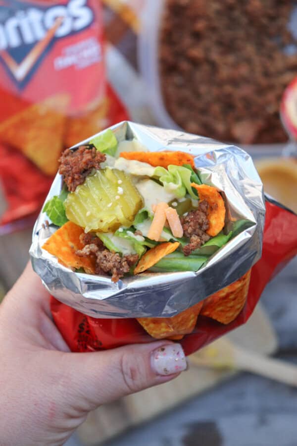 A hand holds an open snack-size Doritos bag filled with taco ingredients, including ground beef, lettuce, shredded cheese, pickles, and chips, creating a walking taco.