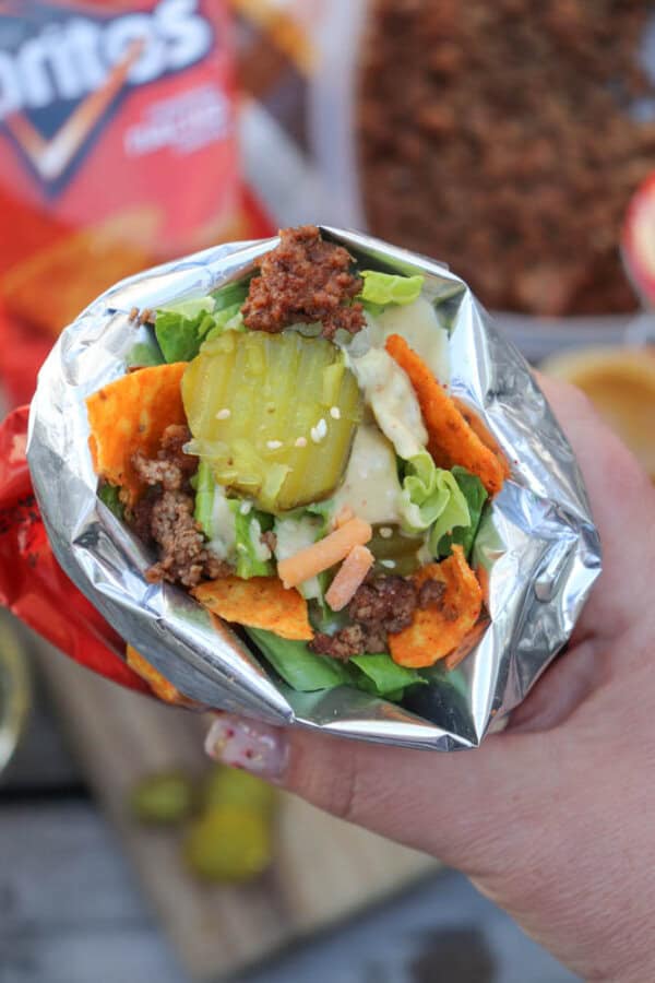 A hand holding a foil-wrapped taco filled with seasoned ground beef, lettuce, shredded cheese, pickles, and Doritos chips, with a blurred bag of Doritos in the background.
