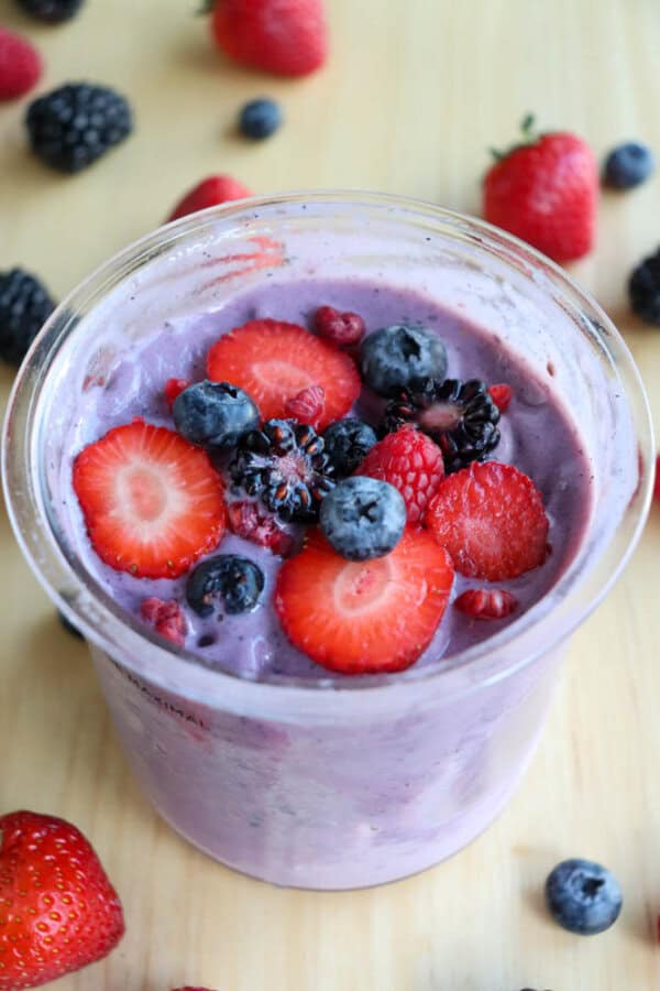 A plastic pint filled with a creamy purple ice cream topped with fresh slices of strawberries, blueberries, raspberries, and blackberries. Additional berries are scattered on the wooden surface around the jar.