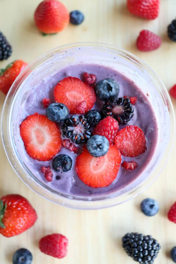 A bowl of berry ice cream topped with sliced strawberries, blackberries, raspberries, and blueberries on a wooden surface. Scattered whole berries surround the bowl.