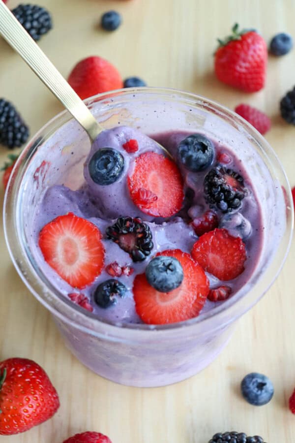A glass cup filled with purple smoothie, topped with sliced strawberries, blueberries, blackberries, and raspberries. A gold spoon is in the cup. Fresh berries are scattered on the wooden surface around the cup.