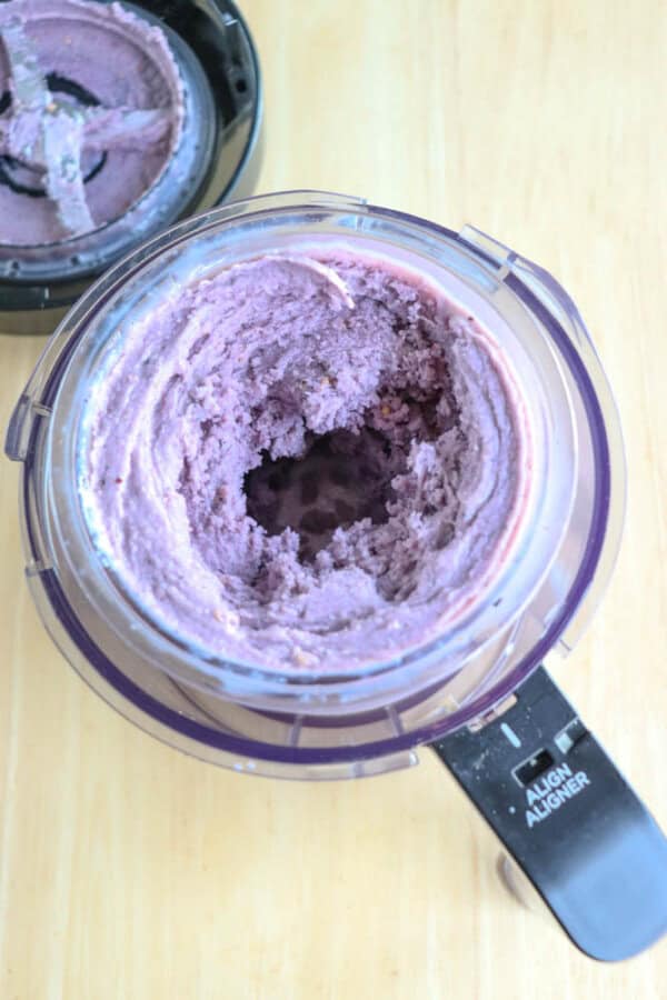 A ninja creami bowl with the lid off contains a thick, creamy purple mixture, possibly a smoothie or acai blend. The container is transparent with measurement markings on the side. It rests on a light wooden surface.