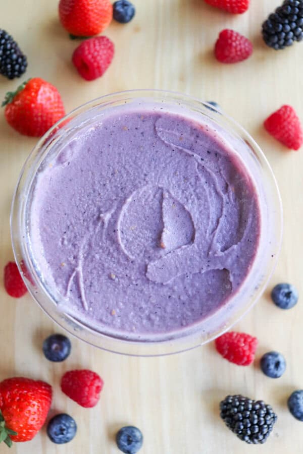 A bowl of smooth purple ice cream on a light wooden table, surrounded by fresh mixed berries, including strawberries, blackberries, raspberries, and blueberries.