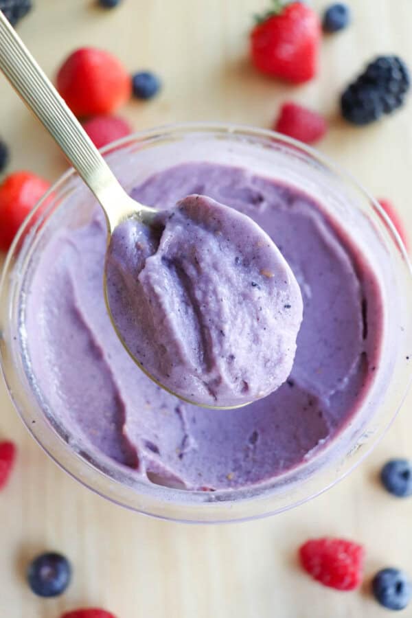 A bowl of creamy purple ice cream with a spoonful held above it. Surrounding the bowl are scattered strawberries, blueberries, raspberries, and blackberries on a light wooden surface.