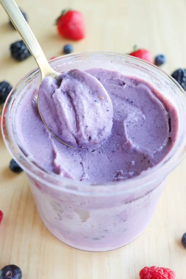 A pint filled with creamy purple ice cream, with a spoon scooping a portion from the top. Fresh berries, including strawberries, raspberries, and blueberries, are scattered around on a light wooden surface.