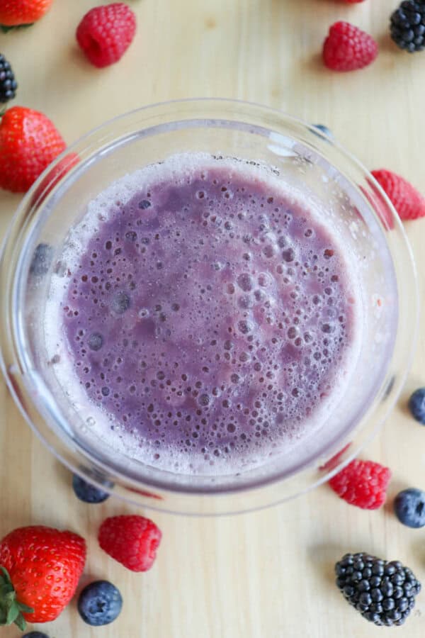 A bowl of frothy purple smoothie sits on a wooden surface, surrounded by fresh berries, including strawberries, raspberries, blueberries, and blackberries. The smoothie is foamy with a slightly bubbly texture on top.