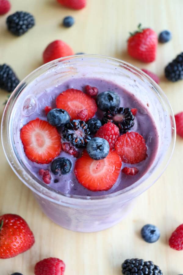 A glass of purple smoothie topped with sliced strawberries, blueberries, raspberries, and blackberries. Surrounding the glass are scattered fresh berries on a wooden surface.