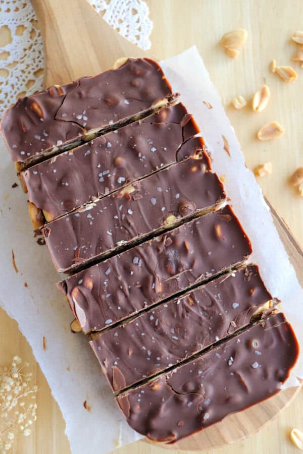Sliced chocolate bars on a wooden board with visible peanuts and a sprinkle of sea salt on top, placed on parchment paper with a doily edge partially visible.