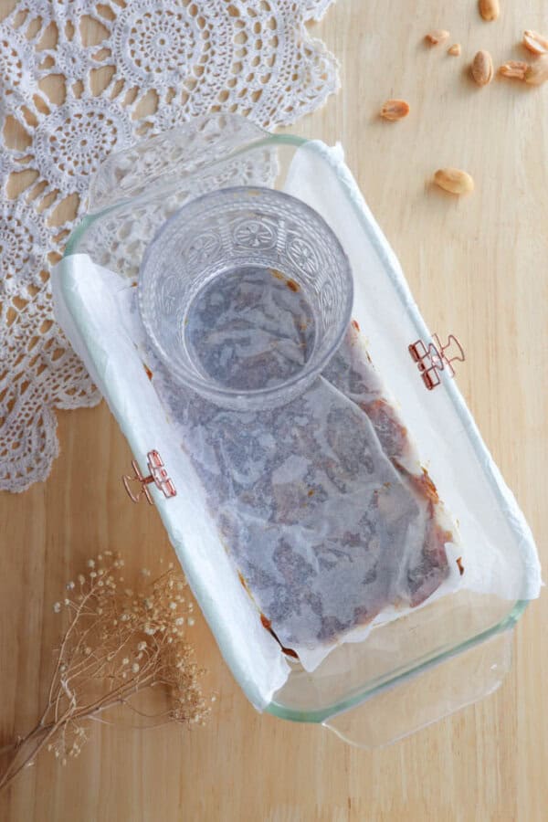 A glass dish lined with parchment paper and weighted with a glass is on a wooden table. Nearby are a few scattered almonds, a piece of lace fabric, and a small bouquet of dried flowers.
