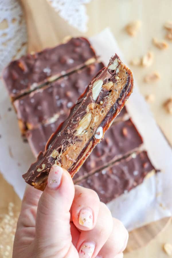 A hand holding a slice of chocolate and nut bar. The bar has visible layers, including caramel, nuts, and a thick chocolate top. More bars are on a wooden surface in the background. The nails are painted with a delicate floral pattern.