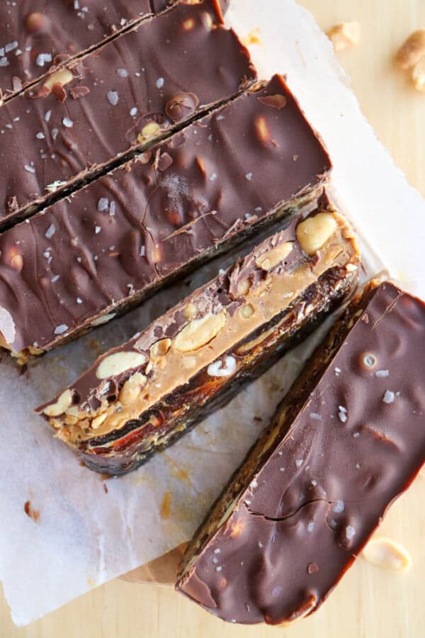 Chocolate-coated bars are sliced on parchment paper, revealing layers of nuts, caramel, and nougat. The surface of the chocolate has a sprinkle of sea salt, and cut sections show a rich, layered texture with visible nuts and caramel.