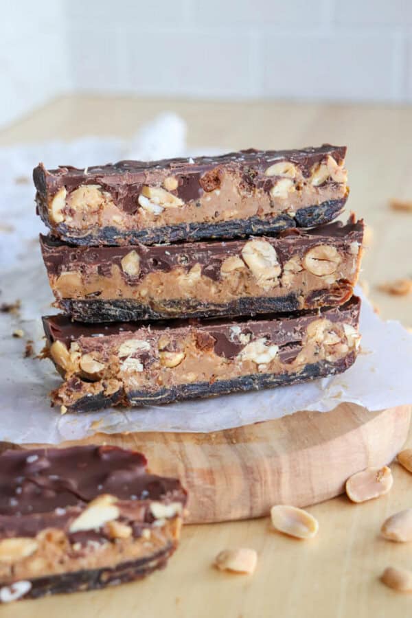 Four stacked chocolate bars on a wooden board. The bars have a chocolate and peanut topping, a peanut butter filling, and a thin dark base. Peanuts are scattered around. The background is a soft beige.