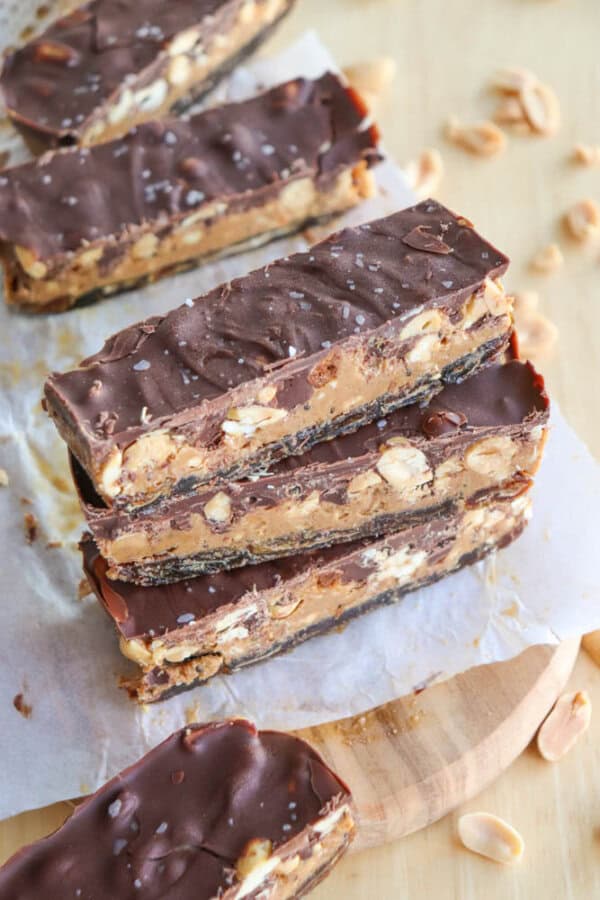 Five chocolate and peanut butter bars stacked on parchment paper, with visible peanuts inside. The top layer is smooth chocolate, sprinkled with sea salt. Crumbled peanuts are scattered around on a wooden surface.