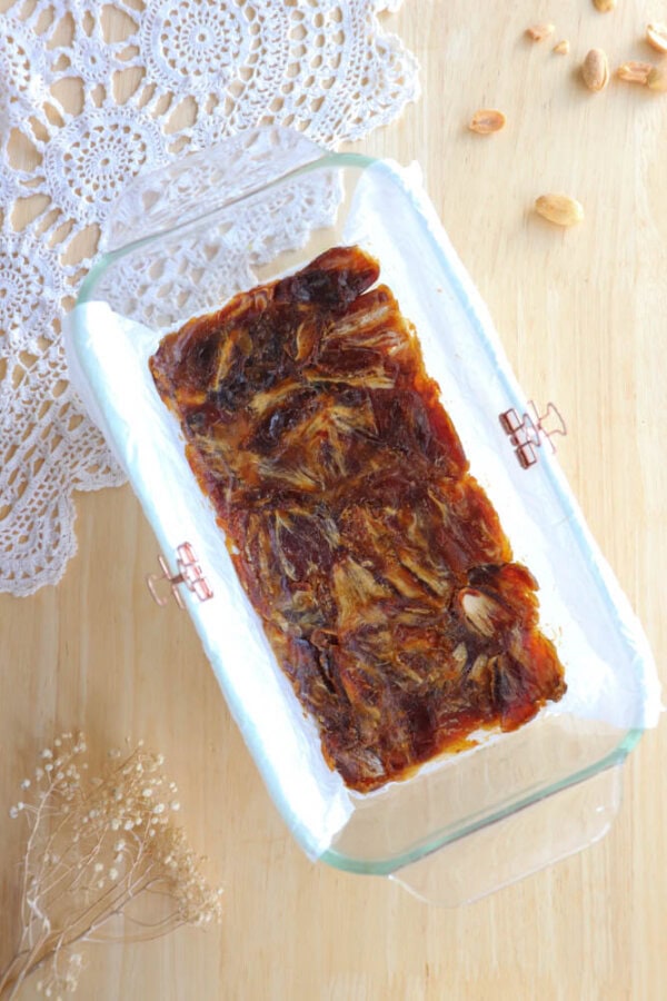 A glass dish contains a rectangular block of date halwa with visible almond pieces. It rests on a wooden surface with a white lace doily and dried flowers nearby. Scattered almond nuts add to the rustic setup.
